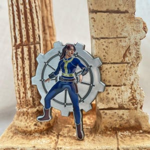 Może przedstawiać: Srebrna metalowa przypinka przedstawiająca karikaturę kobiety w niebieskim kombinezonie stojącej przed dużym kołem zębatym. Kobieta ma brązowe włosy i trzyma pistolet. Przypinka jest kolekcjonerskim przedmiotem z motywem Fallout.