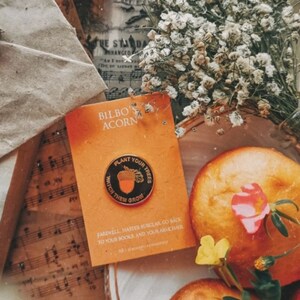 May include: A gold pin with a black background featuring an acorn and the text "Plant Your Trees Watch Them Grow". The pin is on a card with the text "Bilbo Acorn" and a message about going back to books and an armchair.