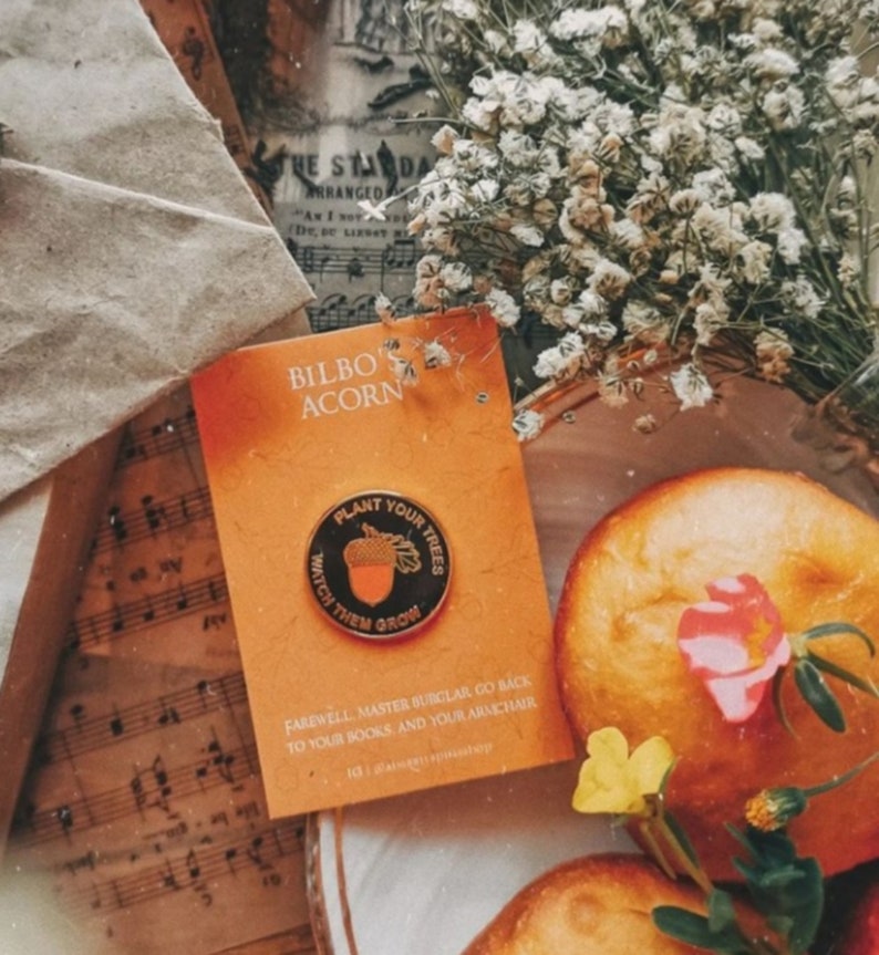 May include: A gold pin with a black background featuring an acorn and the text "Plant Your Trees Watch Them Grow". The pin is on a card with the text "Bilbo Acorn" and a message about going back to books and an armchair.