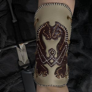 Könnte beinhalten: Ein hellbrauner Lederarmreif mit einem braunen, genähten Design. Der Armreif weist ein detailliertes, symmetrisches Muster auf und ist mit brauner Kordel geschnürt. Die Kanten sind mit weißem Faden genäht. Im Hintergrund ist ein Schwert zu sehen.
