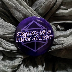 Puede incluir: Botón morado y blanco con un gráfico de dados en blanco y negro y el texto "Crying is a free action".