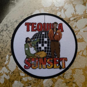 Puede incluir: Un parche redondo con las palabras "TEQUILA SUNSET" en letras rojas y naranjas. El parche presenta dos figuras de dibujos animados, una bola de discoteca y un borde negro. El fondo es blanco.