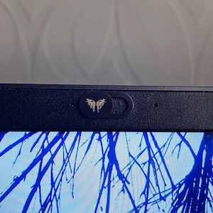 以下が含まれることがあります： 金色の羽根のついたウェブカメラカバーが付いた黒いラップトップ。ラップトップの画面には、青と黒の樹木の枝の画像が表示されています。