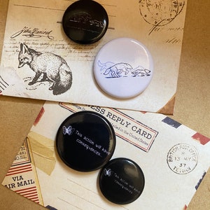 Könnte beinhalten: Vier schwarze und weiße Anstecknadeln mit verschiedenen Designs. Eine Nadel zeigt einen Schmetterling und den Text "This action will have consequences..." Eine andere Nadel zeigt einen Schmetterling und den Text "This action will have consequences". Die dritte Nadel zeigt eine Schwarzweißillustration eines Fuchses. Die vierte Nadel zeigt eine Schwarzweißillustration von drei Hunden.