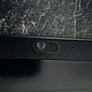 以下が含まれることがあります： 中央に幾何学模様が施された黒いウェブカメラカバー