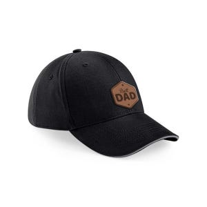 May include: Black baseball cap with a curved peak and a brown leather patch that reads "Best Dad". The cap has a button on top and a grey trim along the peak.