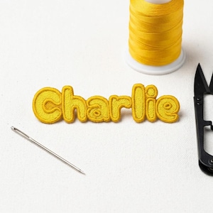 May include: A yellow embroidered patch spelling "Charlie" in a rounded font. A spool of yellow thread, a needle, and black scissors are also present, indicating a crafting project.