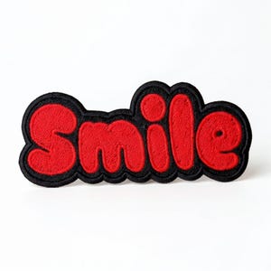 May include: A red embroidered patch featuring the word "Smile" in a rounded font. The letters are red with a black border, set against a white backdrop. The patch is designed for application to clothing or other fabrics.