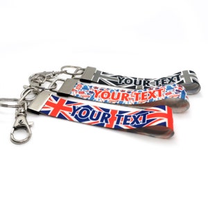 Może przedstawiać: Trzy breloki z srebrnym metalowym kółkiem i paskiem z tkaniny. Paski mają wzór Union Jack z nadrukiem "YOUR TEXT" na każdym.