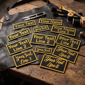 May include: Several black embroidered patches with gold text, including "Your Text" and "Your Name", are displayed. The patches are on a leather surface, alongside sewing tools and thread spools, indicating a craft or personalisation focus.