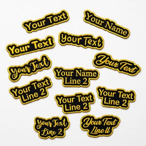 May include: A selection of black embroidered patches with gold lettering and borders. The patches display phrases such as "Your Text" and "Your Name", with some including a second line of text. The patches are arranged on a white background.