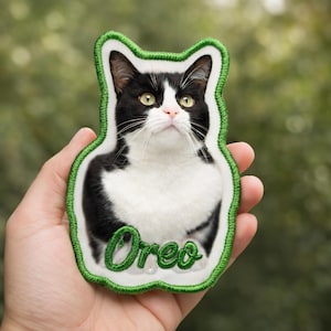 May include: Embroidered patch of a black and white cat with green eyes, named "Oreo" in green script. The patch has a white border with a green stitched outline, ideal for clothing or accessories.