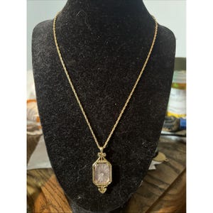 May include: A gold-toned rope chain necklace featuring a rectangular pendant with a beaded metal frame and a central clear, faceted glass stone. The pendant has a sunburst pattern etched into the glass, with small decorative metal flourishes at the top and bottom of the rectangular setting.