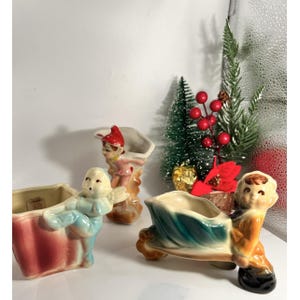 May include: Three vintage ceramic planter figurines. One is a boy pushing a green and brown cart, another is a boy with angel wings, and the third is a boy with a red hat. The planters are in shades of pink, blue, brown, and green.