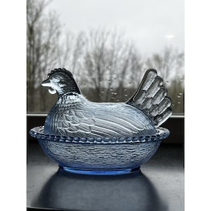 Puede incluir: Un recipiente azul en forma de gallina de vidrio con una superficie texturizada. La gallina tiene patrones detallados de plumas y cresta. El recipiente está sobre una base azul a juego con un borde con cuentas. El fondo muestra una escena borrosa al aire libre.