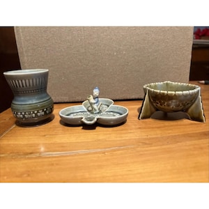 Puede incluir: Tres piezas decorativas de cerámica sobre una superficie de madera. Los artículos incluyen un pequeño jarrón con tonos azules y verdes, un plato en forma de trébol con una figura y un cuenco con un diseño único y detalles en color beige.