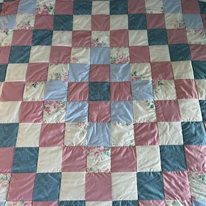 Pode incluir: Um cobertor de retalhos com um padrão de tabuleiro de xadrez em tons de rosa, azul e creme. O cobertor é feito de vários tecidos florais e tem um toque rústico e vintage.