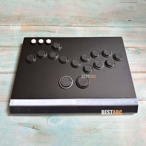 Op de afbeelding: Zwarte arcade game controller met meerdere knoppen en een joystick. De controller heeft een rechthoekige vorm met de tekst "BESTABC" onderaan. De knoppen zijn zwart en wit.