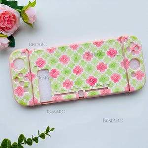 May include: A protective gaming console case featuring a repeating pattern of pink cherry blossoms and light green four-leaf clovers on a pale background. The case includes cutouts for buttons and joysticks, with the text BestABC printed on it.