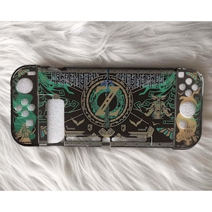 Może przedstawiać: Czarny i zielony pokrowiec ochronny na konsolę do gier. Etui ma szczegółowy wzór z centralnym emblematem, stylizowaną grafiką i mieczem. Projekt jest podkreślony zielonymi i złotymi akcentami.