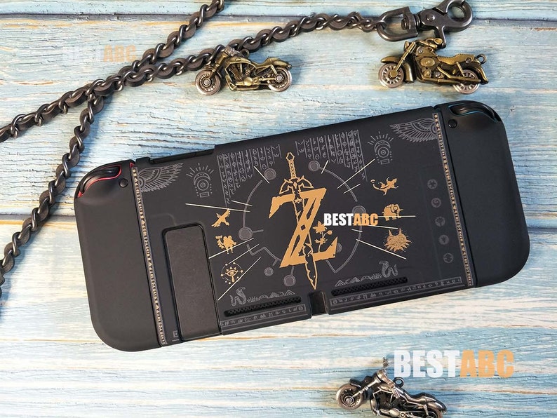 K&ouml;nnte beinhalten: Schwarze Nintendo Switch-H&uuml;lle mit goldenem Zelda-Design. Das Design zeigt ein Schwert, einen Schild und andere Symbole aus dem Spiel. Die H&uuml;lle ist aus strapazierf&auml;higem Kunststoff und hat ein weiches Innenfutter zum Schutz Ihrer Konsole.