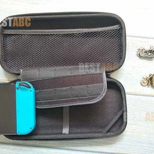 K&ouml;nnte beinhalten: Eine schwarze Tragetasche mit Rei&szlig;verschluss und einem Netzfach im Inneren. Die Tasche ist ge&ouml;ffnet und eine blau-schwarze Nintendo Switch-Konsole befindet sich darin. Die Tasche liegt auf einer hellblauen Oberfl&auml;che.
