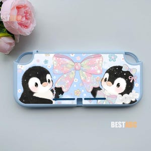 May include: A light blue protective console case featuring a graphic of two black and white penguins holding a large, iridescent, multi-colored bow. The design includes small flower accents and sparkles against a light blue background.