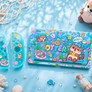 May include: A light blue protective case for a gaming console and two matching controller covers, featuring a colorful underwater pattern with cartoon otters, jellyfish, fish, and the word OTTER in bold, multi-colored bubble letters.