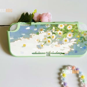 May include: A light green protective console case featuring a printed design of white daisies and green foliage against a light blue and white background. The text A beautiful day is printed on the front. A pastel-colored beaded bracelet is nearby.