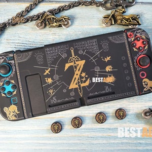 Puede incluir: Consola Nintendo Switch negra con un diseño personalizado de temática Zelda. El diseño presenta una espada dorada, un símbolo dorado de la Triforce y otras imágenes de temática Zelda. La consola tiene botones azules y rojos y un borde negro y dorado.