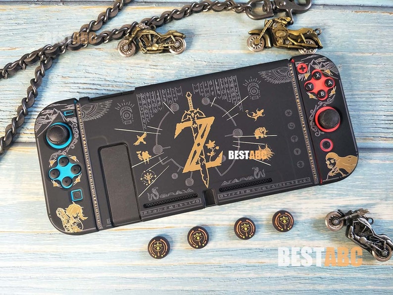 K&ouml;nnte beinhalten: Schwarze Nintendo Switch-Konsole mit einem benutzerdefinierten Zelda-Design. Das Design zeigt ein goldenes Schwert, ein goldenes Triforce-Symbol und andere Zelda-bezogene Bilder. Die Konsole hat blaue und rote Kn&ouml;pfe und eine schwarze und goldene Einfassung.