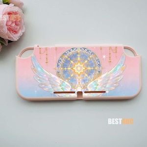 Könnte beinhalten: Eine hellrosa Schutzhülle für eine Konsole mit einem zentralen blauen und goldenen kreisförmigen Himmels-Emblem, das von irisierenden weißen Flügeln flankiert wird. Das Design zeigt hängende Goldketten auf einem rosa-blauen Farbverlauf.