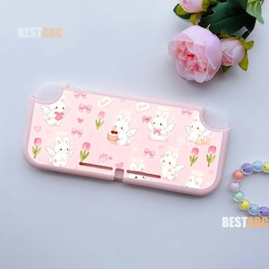 Könnte beinhalten: Eine hellrosa Schutzhülle für eine Konsole mit einem sich wiederholenden Muster aus weißen geflügelten Hasen, rosa Tulpen, kleinen Herzen und Schleifen. Der Text BESTABC ist oben links und unten rechts aufgedruckt. Die Hülle hat Aussparungen.