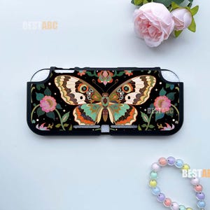 May include: A black protective gaming console case featuring a central butterfly graphic with intricate patterns in shades of orange, teal, and cream, surrounded by pink floral motifs and small white stars against a dark background.