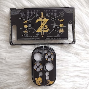 以下が含まれることがあります： ゼルダをテーマにした、黒と金のNintendo Switchアクセサリー。上部には金の剣と文字「Z」があります。下部には銀と金のアクセントがあります。アクセサリーは白くふわふわした表面にあります。
