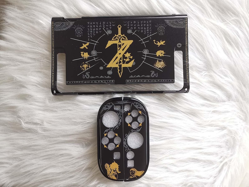 以下が含まれることがあります： ゼルダをテーマにした、黒と金のNintendo Switchアクセサリー。上部には金の剣と文字「Z」があります。下部には銀と金のアクセントがあります。アクセサリーは白くふわふわした表面にあります。