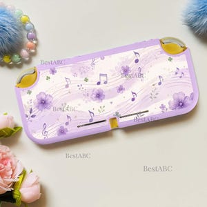 May include: A handheld gaming console with a light purple shell featuring a pattern of purple flowers, musical notes, and wavy lines. The device has yellow joystick caps and the text BestABC is printed multiple times across the surface.