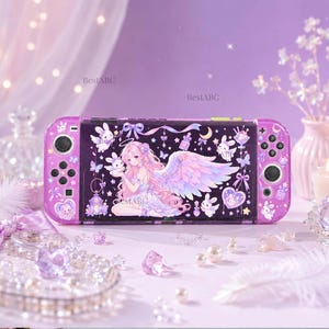 Puede incluir: Una consola de juegos portátil de color morado con un gráfico de un ángel de pelo largo rosa y alas, rodeado de pequeños conejos blancos, estrellas y lazos. El texto BestABC está impreso en la pantalla. El dispositivo tiene joysticks negros.