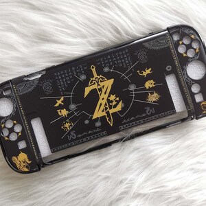 以下が含まれることがあります： 黒色のNintendo Switchケース。金色のゼルダのデザインで、ロゴ、剣、様々なシンボルが描かれています。ボタンやポート用の切り欠きがあり、白くふわふわした背景に配置されています。