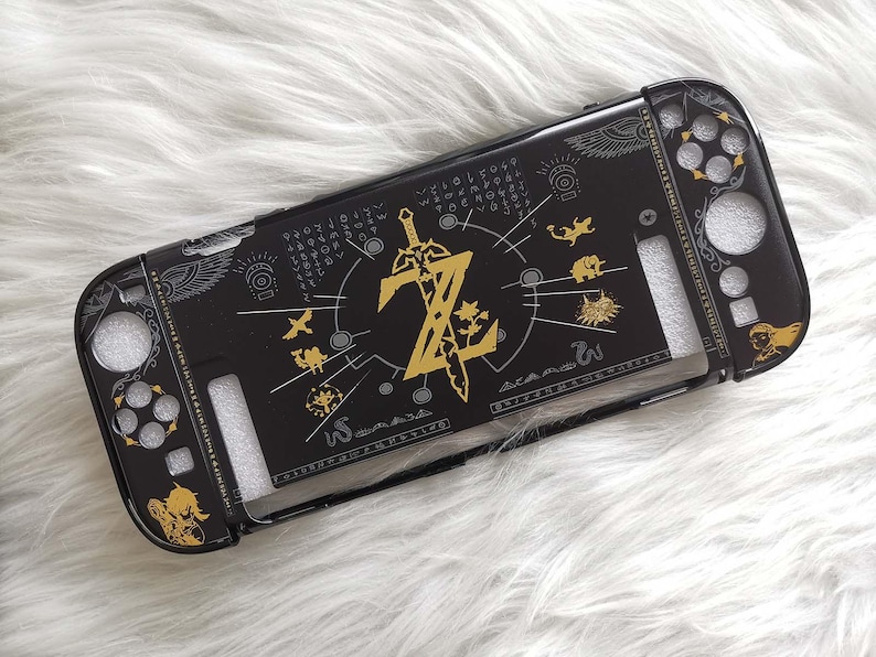 以下が含まれることがあります： 黒色のNintendo Switchケース。金色のゼルダのデザインで、ロゴ、剣、様々なシンボルが描かれています。ボタンやポート用の切り欠きがあり、白くふわふわした背景に配置されています。