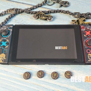 K&ouml;nnte beinhalten: Eine schwarze Nintendo Switch-Konsole mit benutzerdefinierten Frontplatten in Gold und Schwarz. Die Konsole hat einen schwarzen Bildschirm und ist von einer Kette mit Motorrad-Anh&auml;ngern umgeben. Die Konsole befindet sich auf einer hellblauen Holzoberfl&auml;che mit vier kleinen, runden, goldenen und schwarzen Aufklebern.