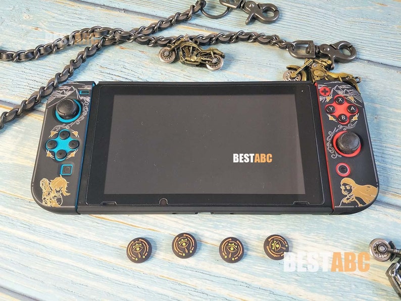 K&ouml;nnte beinhalten: Eine schwarze Nintendo Switch-Konsole mit benutzerdefinierten Frontplatten in Gold und Schwarz. Die Konsole hat einen schwarzen Bildschirm und ist von einer Kette mit Motorrad-Anh&auml;ngern umgeben. Die Konsole befindet sich auf einer hellblauen Holzoberfl&auml;che mit vier kleinen, runden, goldenen und schwarzen Aufklebern.