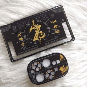 以下が含まれることがあります： 黒と金のNintendo Switchケースとコントローラーカバー。ケースは、文字「Z」と剣、その他のシンボルを備えた金色のデザインが特徴です。コントローラーカバーは、お揃いのデザインです。