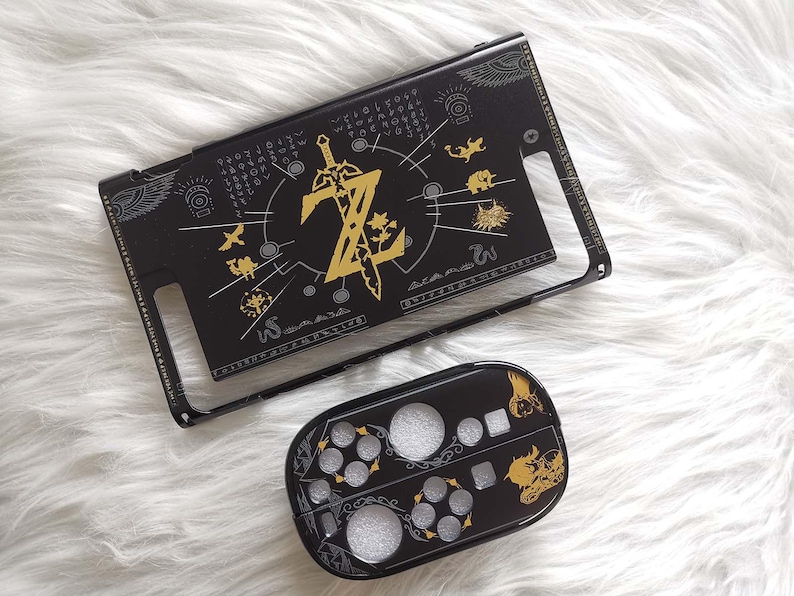 以下が含まれることがあります： 黒と金のNintendo Switchケースとコントローラーカバー。ケースは、文字「Z」と剣、その他のシンボルを備えた金色のデザインが特徴です。コントローラーカバーは、お揃いのデザインです。