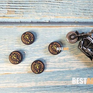 K&ouml;nnte beinhalten: Vier schwarze und goldene runde Controller-Kappen mit einem stilisierten Augen-Design. Die Kappen liegen auf einer blauen Holzoberfl&auml;che mit einem kleinen Spielzeugmotorrad.