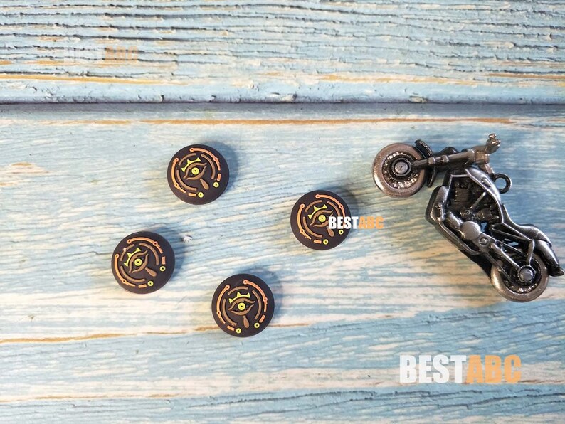 K&ouml;nnte beinhalten: Vier schwarze und goldene runde Controller-Kappen mit einem stilisierten Augen-Design. Die Kappen liegen auf einer blauen Holzoberfl&auml;che mit einem kleinen Spielzeugmotorrad.
