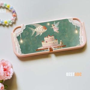 May include: A light pink protective console case featuring a green background with a white polka-dot fish, a small fairy, and a tiered strawberry cake. The case includes white lace-like trim and hanging light bulb graphics. A pastel bead wrist strap is attached.