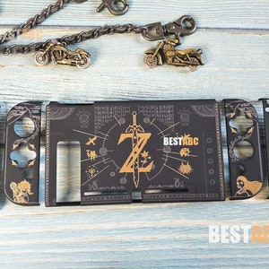 K&ouml;nnte beinhalten: Eine schwarze Nintendo Switch-H&uuml;lle mit einem goldenen Zelda-Design. Die H&uuml;lle zeigt ein stilisiertes Bild des Meisterschwerts und der Triforce, umgeben von komplizierten Mustern und Symbolen. Der Text "BESTABC" ist auf der H&uuml;lle aufgedruckt.