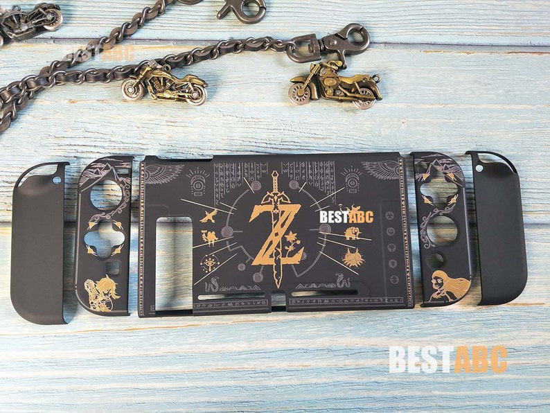 K&ouml;nnte beinhalten: Eine schwarze Nintendo Switch-H&uuml;lle mit einem goldenen Zelda-Design. Die H&uuml;lle zeigt ein stilisiertes Bild des Meisterschwerts und der Triforce, umgeben von komplizierten Mustern und Symbolen. Der Text "BESTABC" ist auf der H&uuml;lle aufgedruckt.