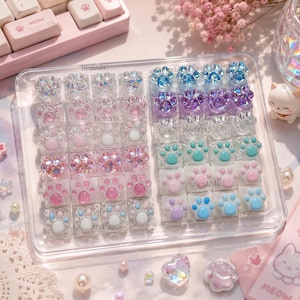 Op de afbeelding: Een verzameling vierkante toetsenbordtoetsen in een doorzichtige plastic tray, elk met een verhoogd kattenpootontwerp. De toetsen hebben verschillende afwerkingen, waaronder glitterende, doorschijnende en effen pastelkleuren zoals roze, blauw en paars.